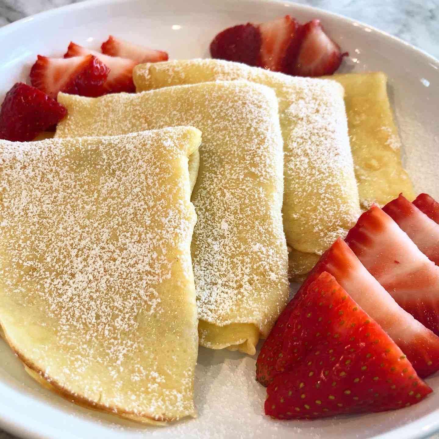 a plate of four crepes folded into quarters, dusted with powdered sugar with sliced strawberries on the side