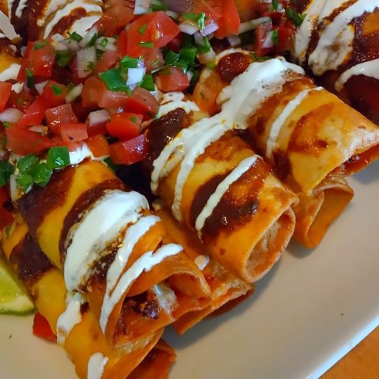 a close up look at a pile of golden spicy chicken taquitos topped with sour cream and fresh pico de gallo.