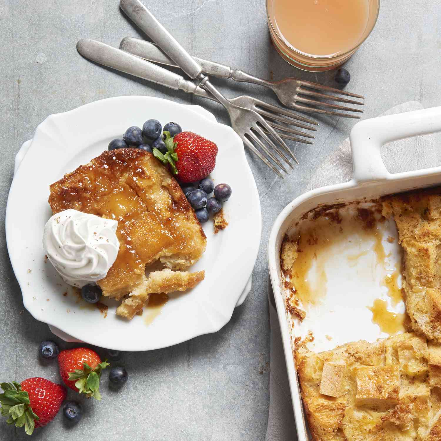 image of french toast with berries and whipped cream on a plate, and french toast in a baking dish, with forks and a glass of juice on the side
