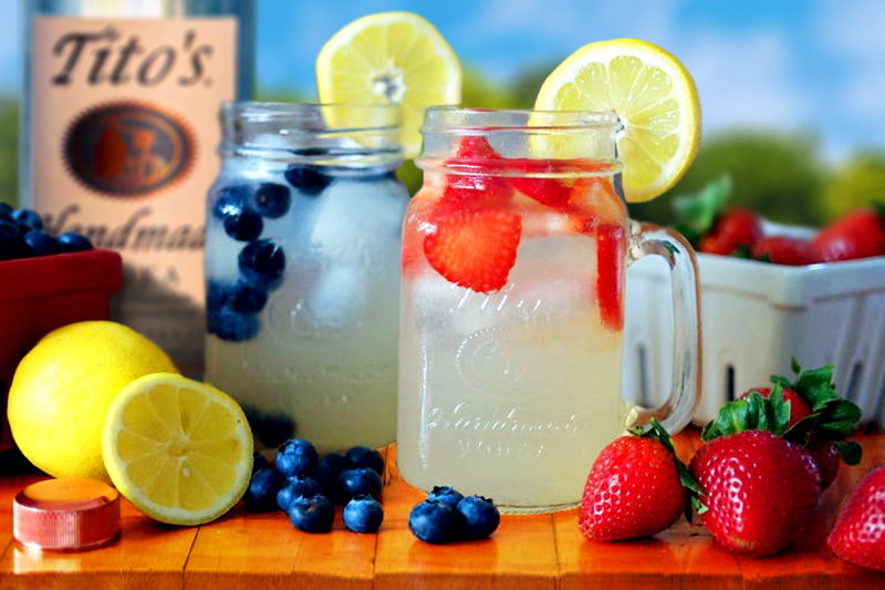 two mason jar glasses of spiked lemonade, one with blueberries, one with strawberries, both garnished with a slice of lemon