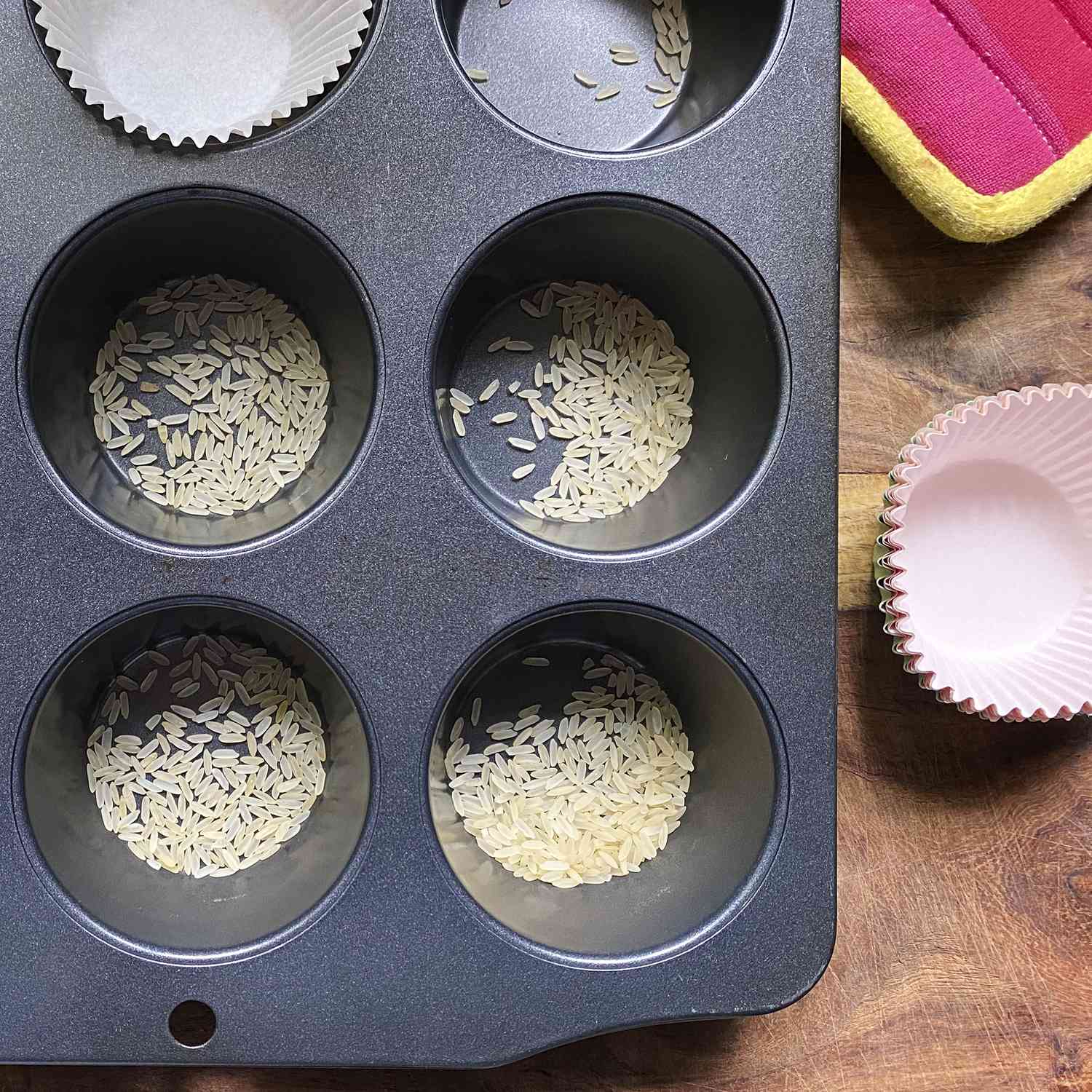 Empty muffin tin with dry rice scattered in the bottom of each muffin cup. Some liners sitting next to the muffin tin to go in on top of the rice.