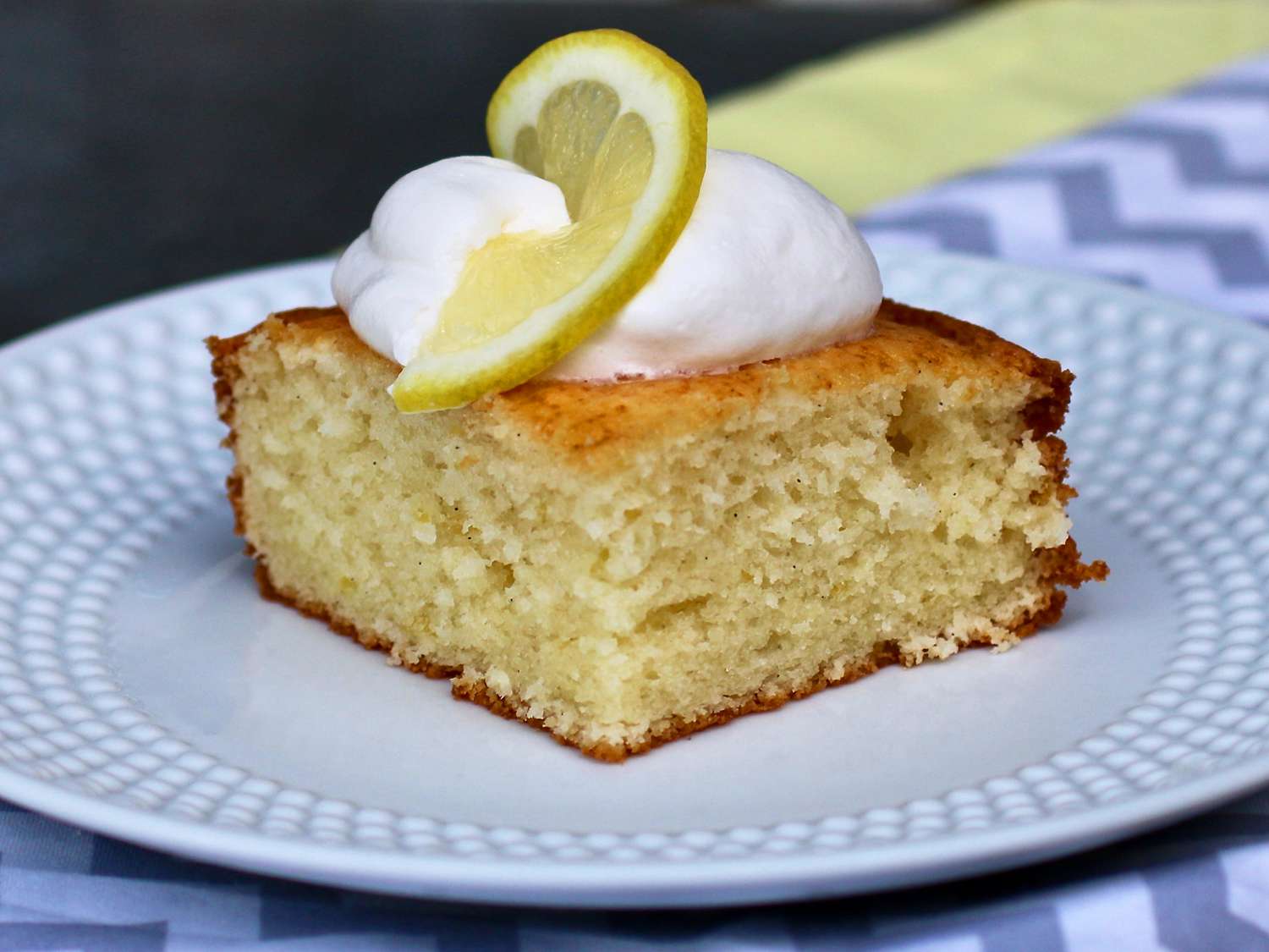 close up view of a slice of Simple Lemon Cake garnished with whipped cream and a slice of lemon on a white plate