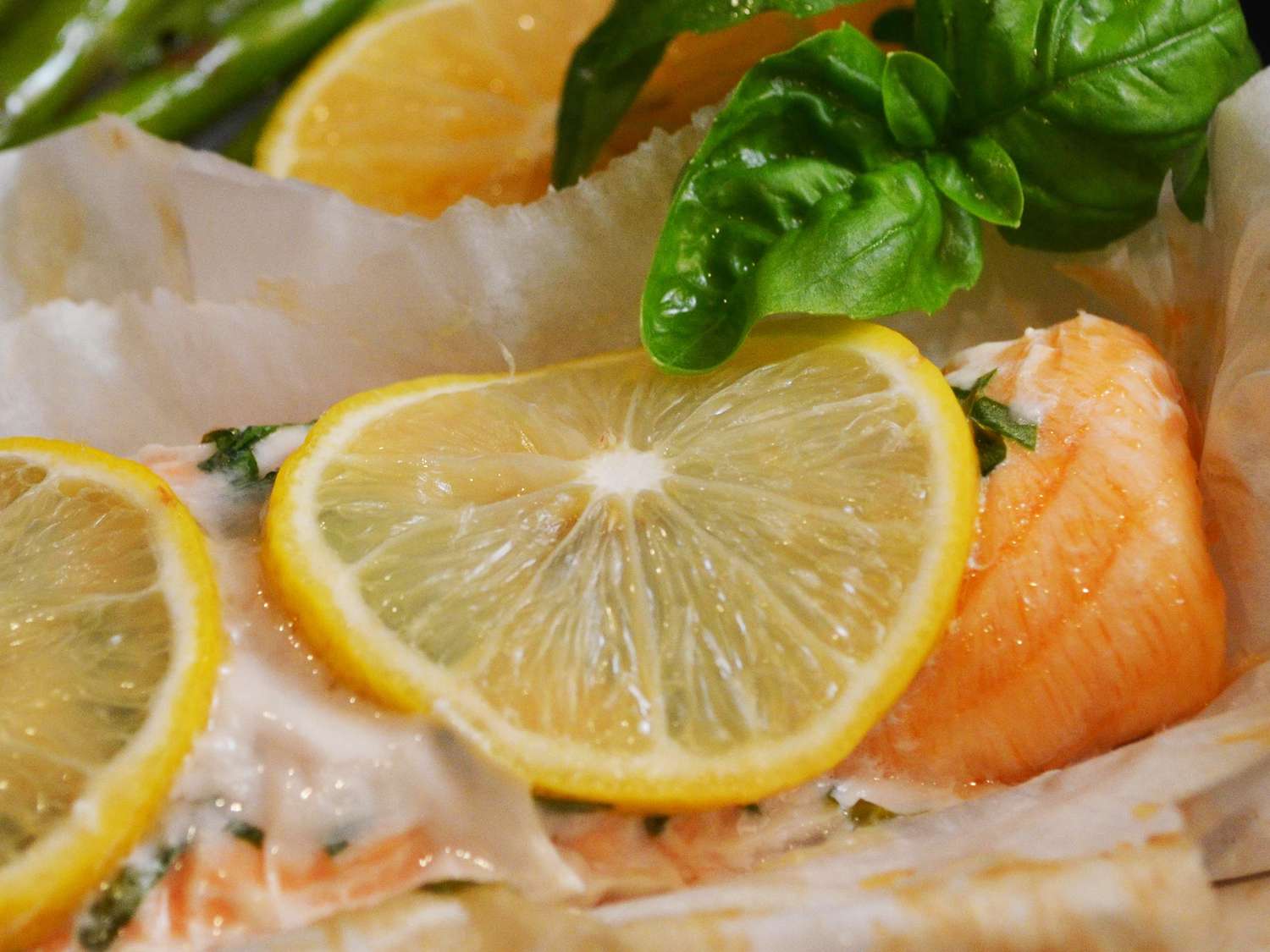 close up view of a salmon fillet with a lemon slice and herbs on parchment paper, with fresh basil leaves