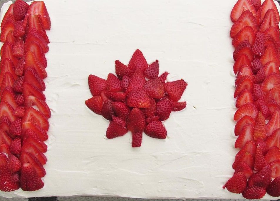 top-down view of a rectangular cake with white frosting and red panels and maple leaf design made out of sliced strawberries