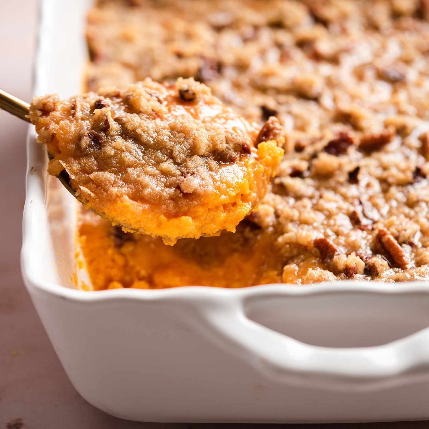 a close up view of a spoonful of sweet potato casserole, topped with pecans and brown sugar, being lifted from a white baking dish.
