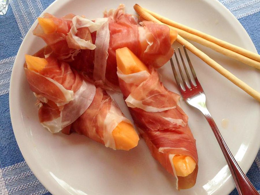 close up view of Prosciutto e Melone (Italian Ham and Melon) on a white plate with bread sticks and a fork