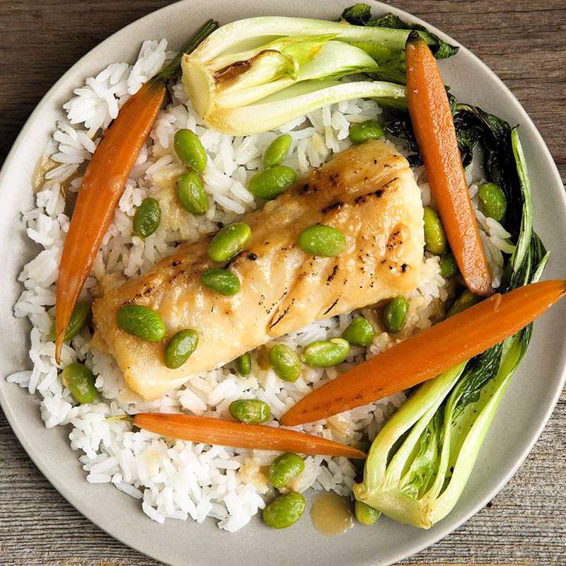 close up view of a Miso-Glazed Black Cod fillet served with white rice, carrots, bok choy and edamame on a plate