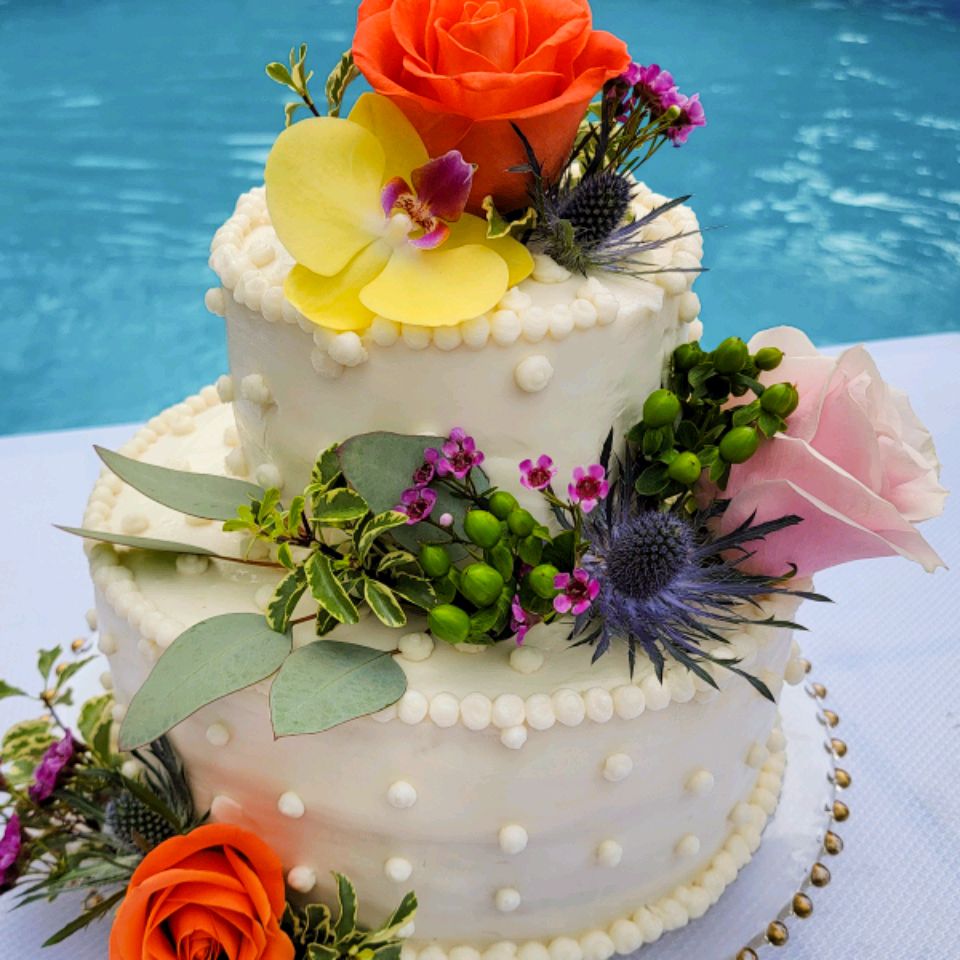 close up view of White Almond Wedding Cake garnished with white icing and fresh flowers with a swimming pool in the background