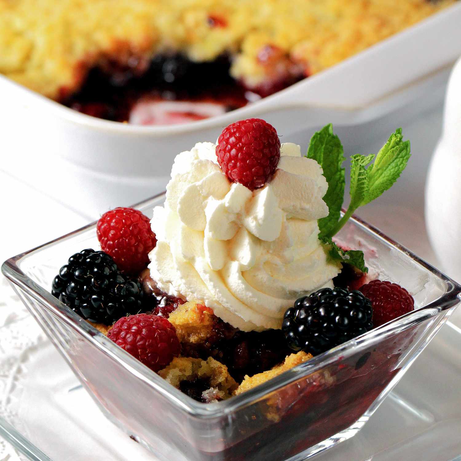 close up view of a Triple Berry Crisp served in a glass bowl with cream and fresh mint, also Triple Berry Crisp in a white baking dish