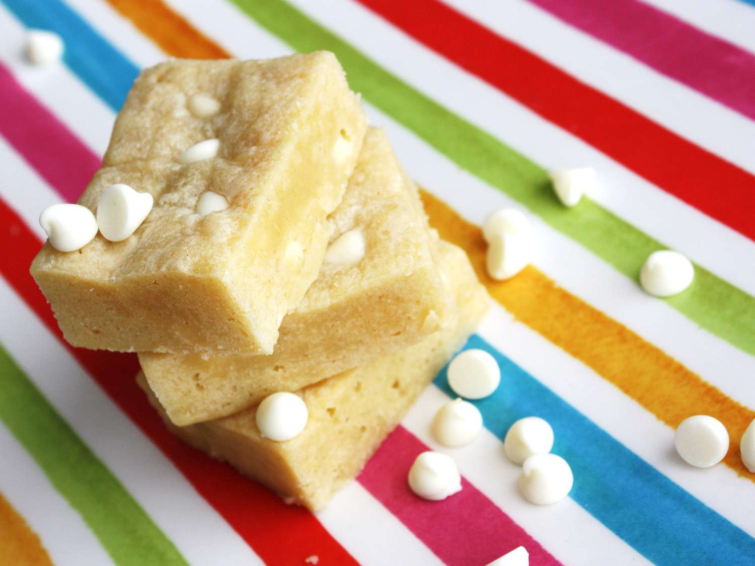 close up view of a stack of White Chocolate Blondies on a rainbow surface with white chocolate chips