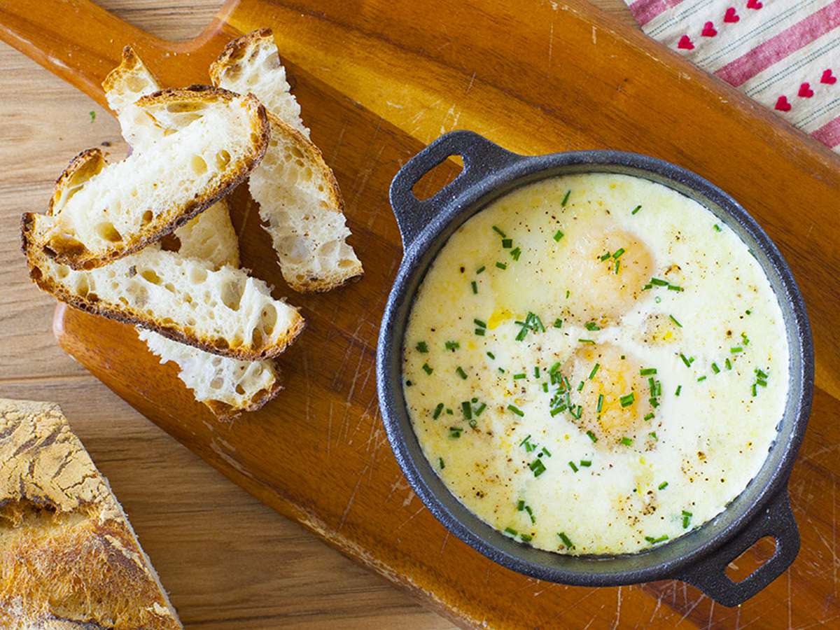 close up view of a Oeufs Cocotte (Baked Eggs) in a small pot on a wooden cutting boar, with bread slices