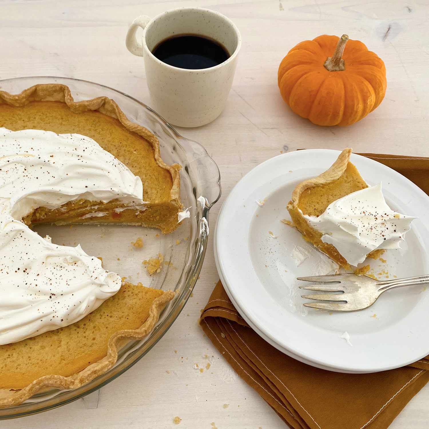 close up view of a Sweet Potato Pie with cream in a pie pan, a slice of Sweet Potato Pie on a white plate with a fork, a cup with coffee and a small pumpkin