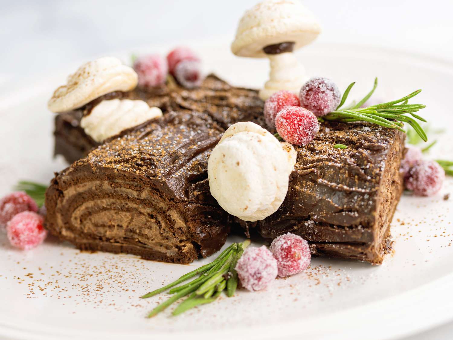 a low angle, close up view of a chocolate yule log topped with meringue mushrooms, sugared cranberries, and rosemary sprigs.