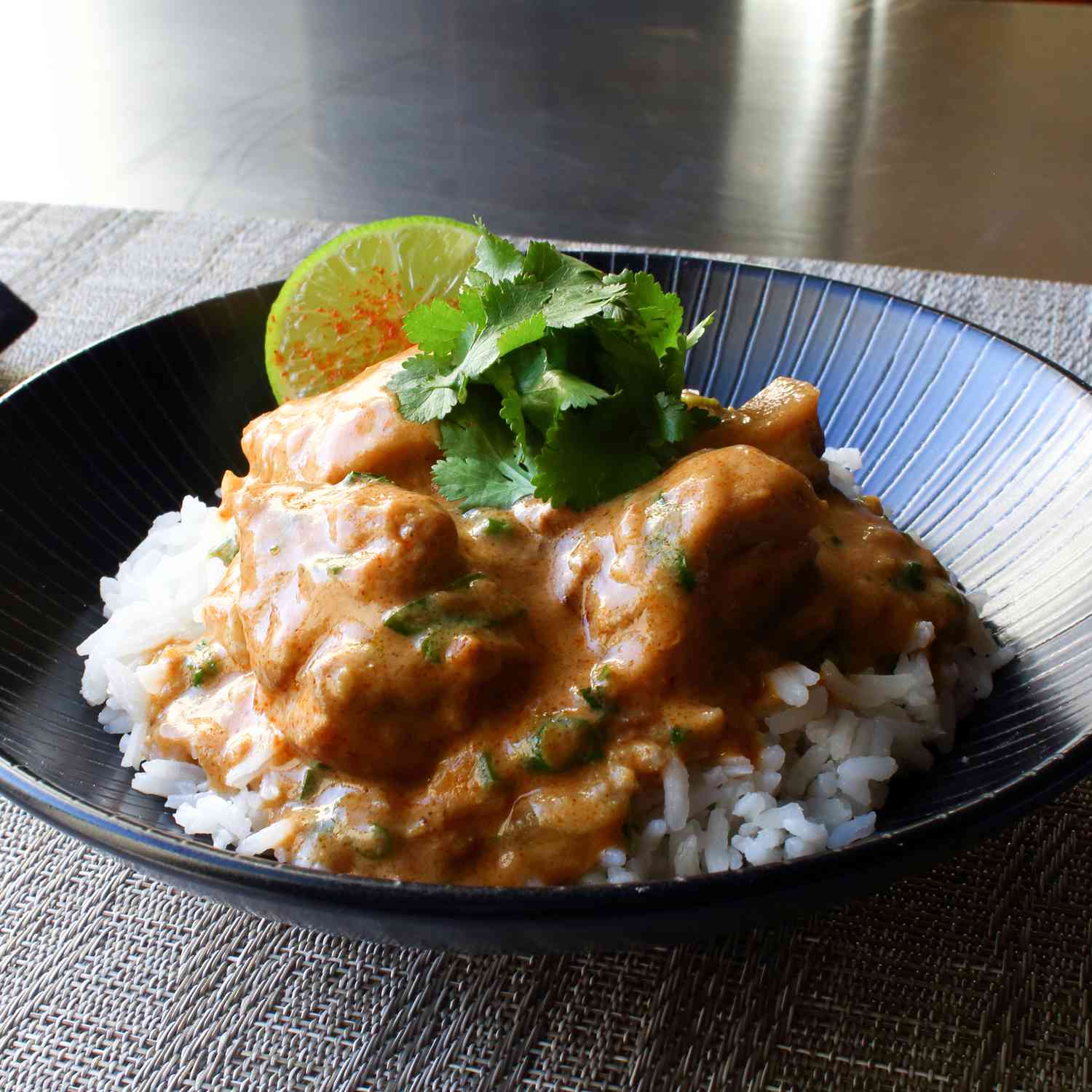 close up view of Creamy Cashew Chicken Curry over white rice, garnished with fresh herbs and a lime wedge in a black bowl