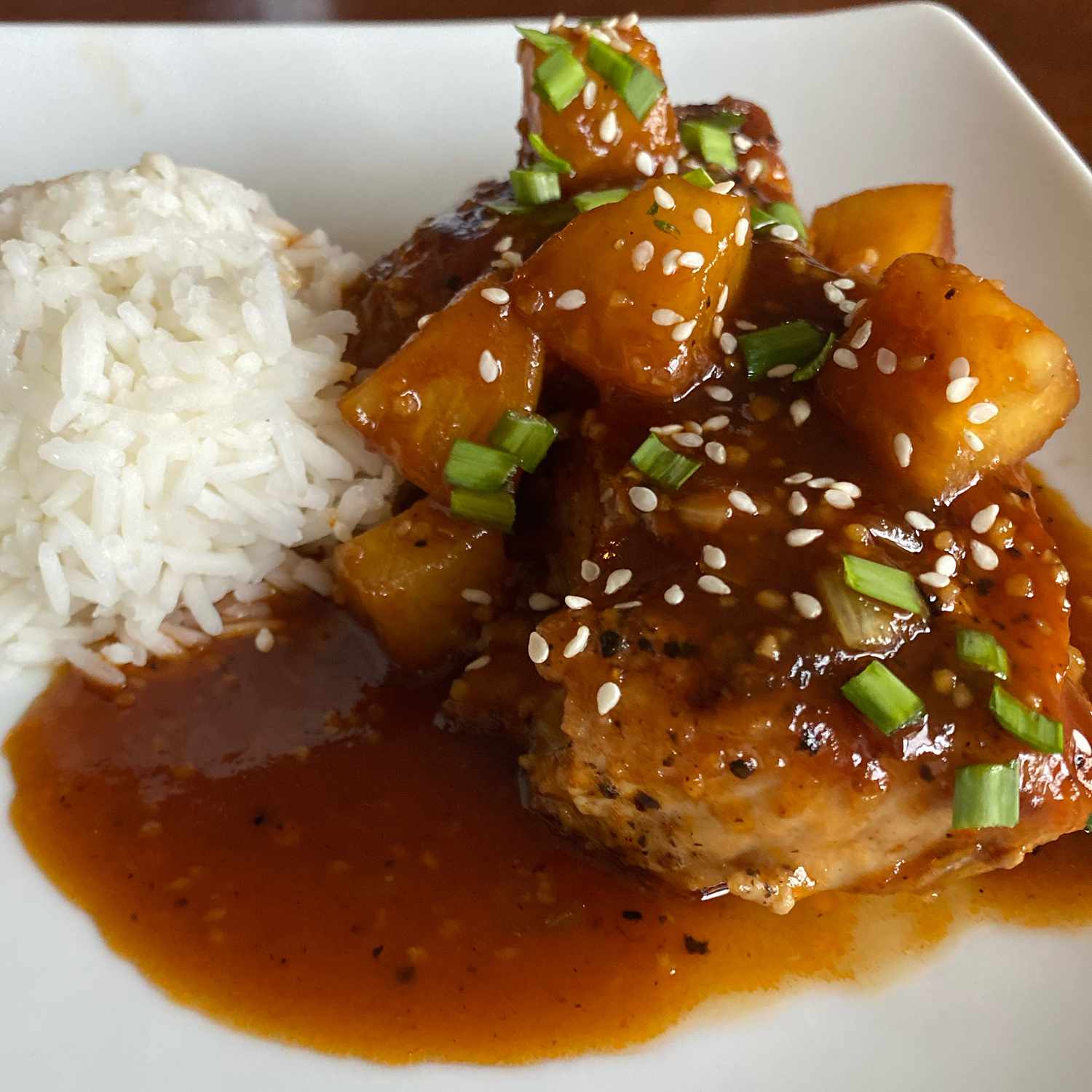 close up view of Sweet and Sour Pork Tenderloin with pineapple piece, green onions, and sesame seeds, served with white rice on a white plate