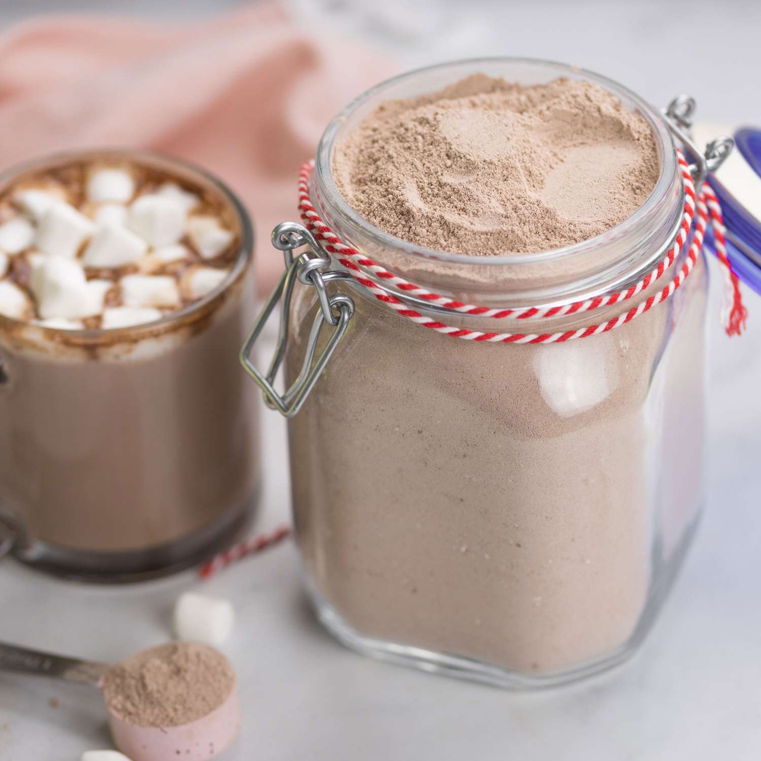 a low angle close up view of a jar of hot coco mix with a single cup of hot coco topped with marshmallows sitting next to it.