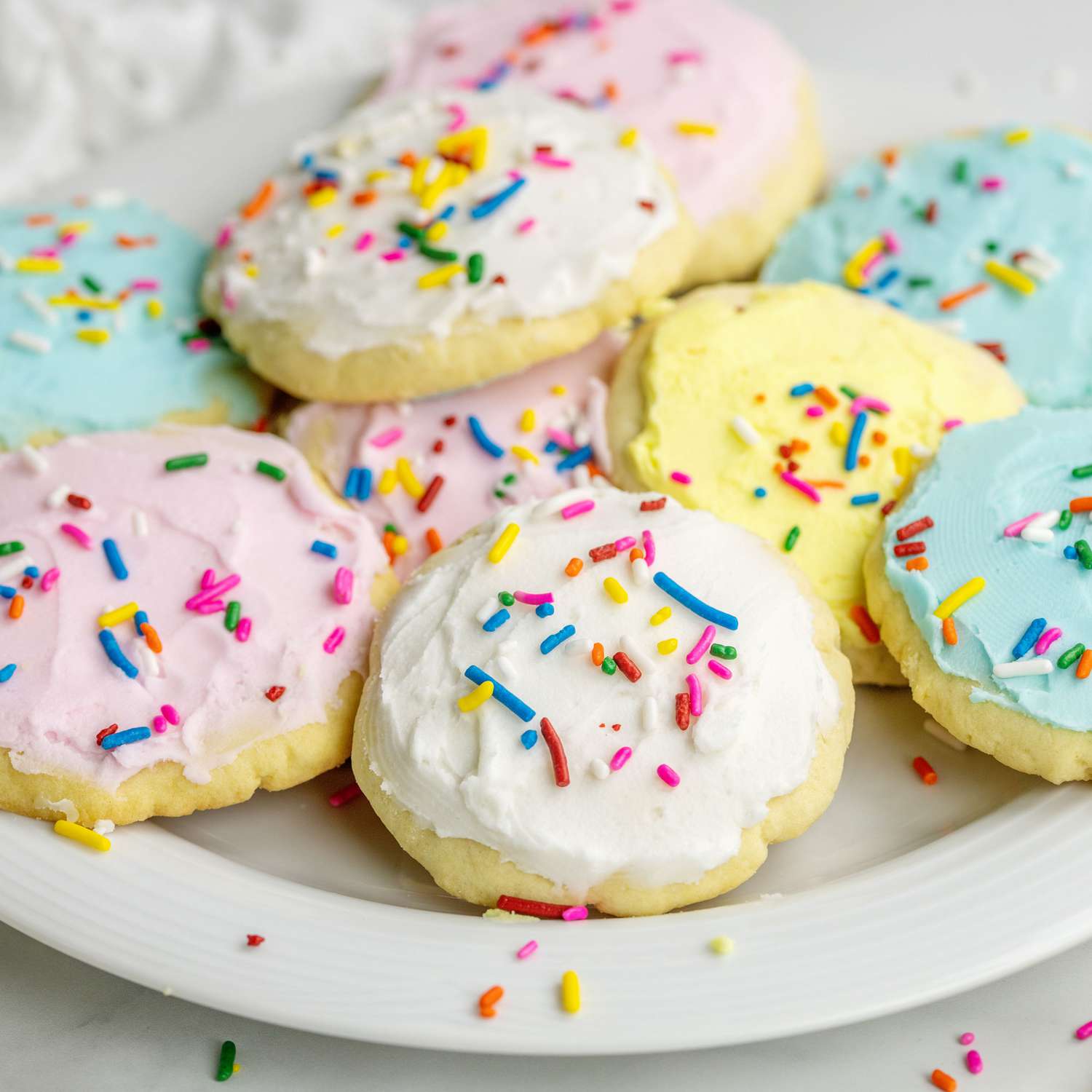 a low angle close up view of a plate full of pastel frosted sugar cookies topped with rainbow colored sprinkles.