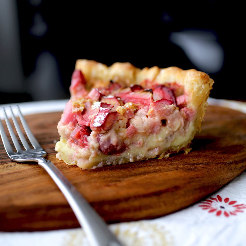 close up view of a slice of Strawberry Rhubarb Custard Pie next to a spoon on a wooden cutting board