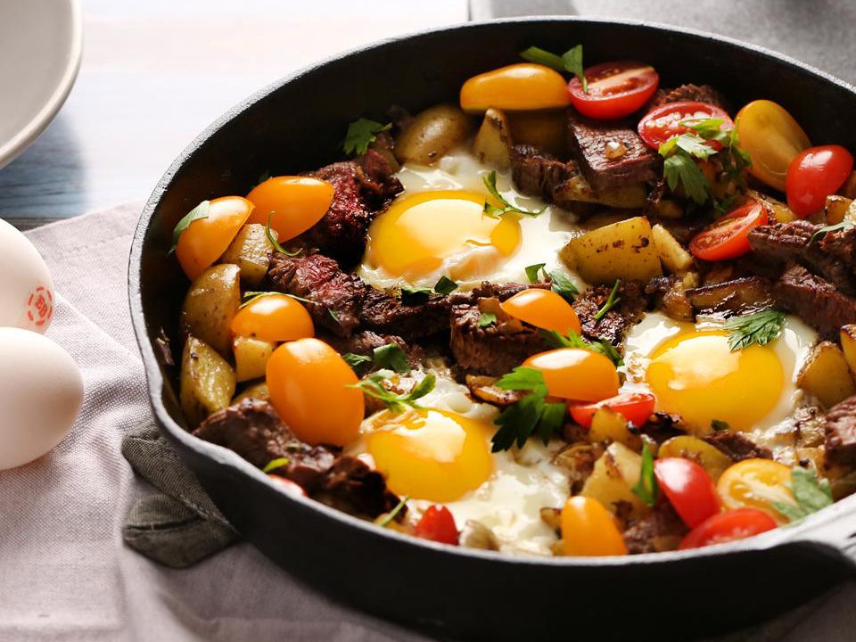 close up view of a Steak and Egg Hash garnished with tomatoes and fresh herbs in a cast iron skillet