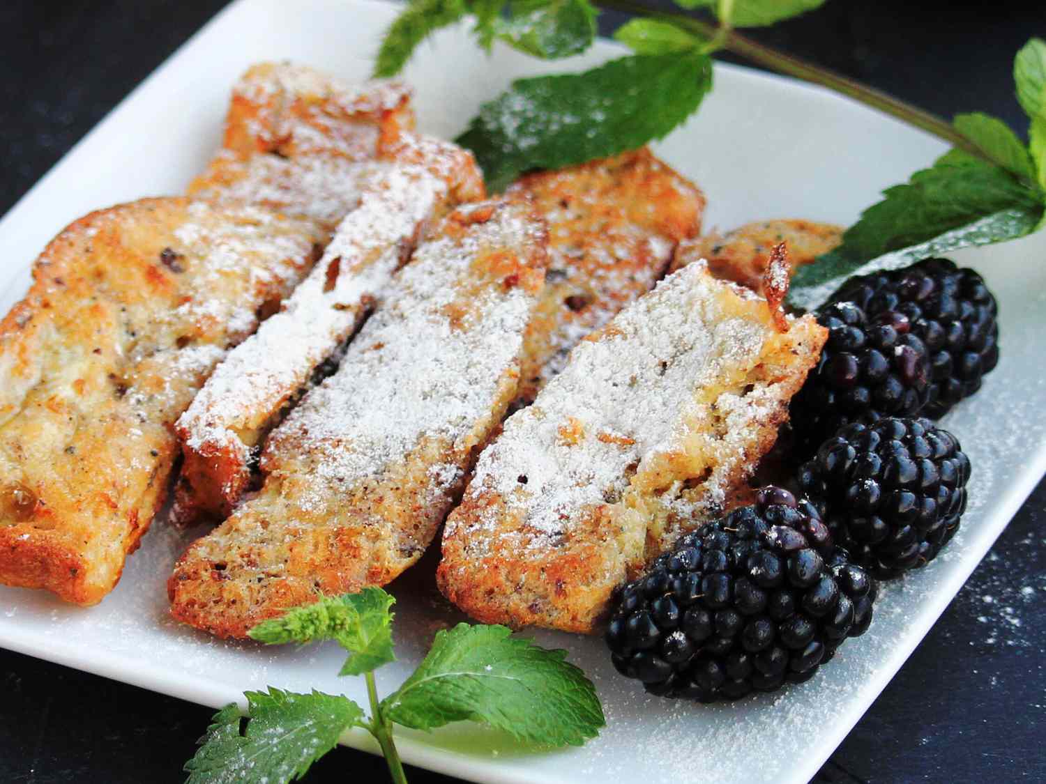 close up view of French Toast Sticks garnished with powdered sugar, served with blackberries and fresh mint on a white plate