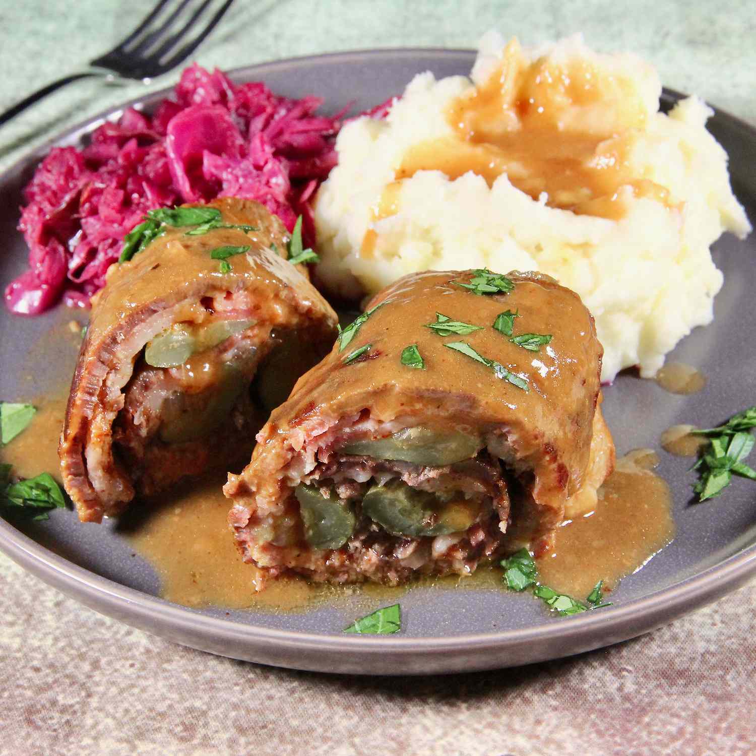 close up view of a Beef Rouladen garnished with gravy and fresh herbs, served on a plate with red cabbage and mashed potatoes with gravy