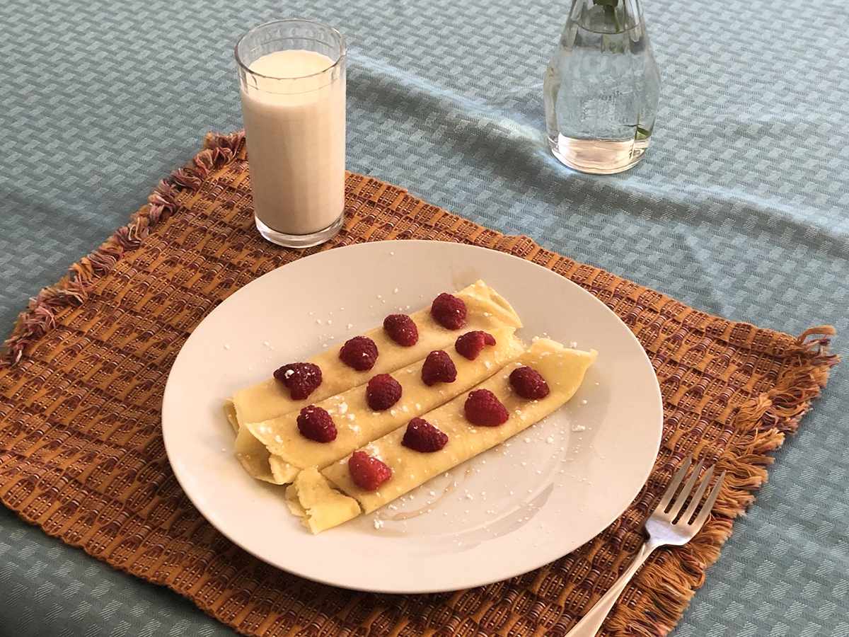 close up view of Fluffy Swedish Pancakes garnished with raspberries and powdered sugar on a white plate, served a glass of milk