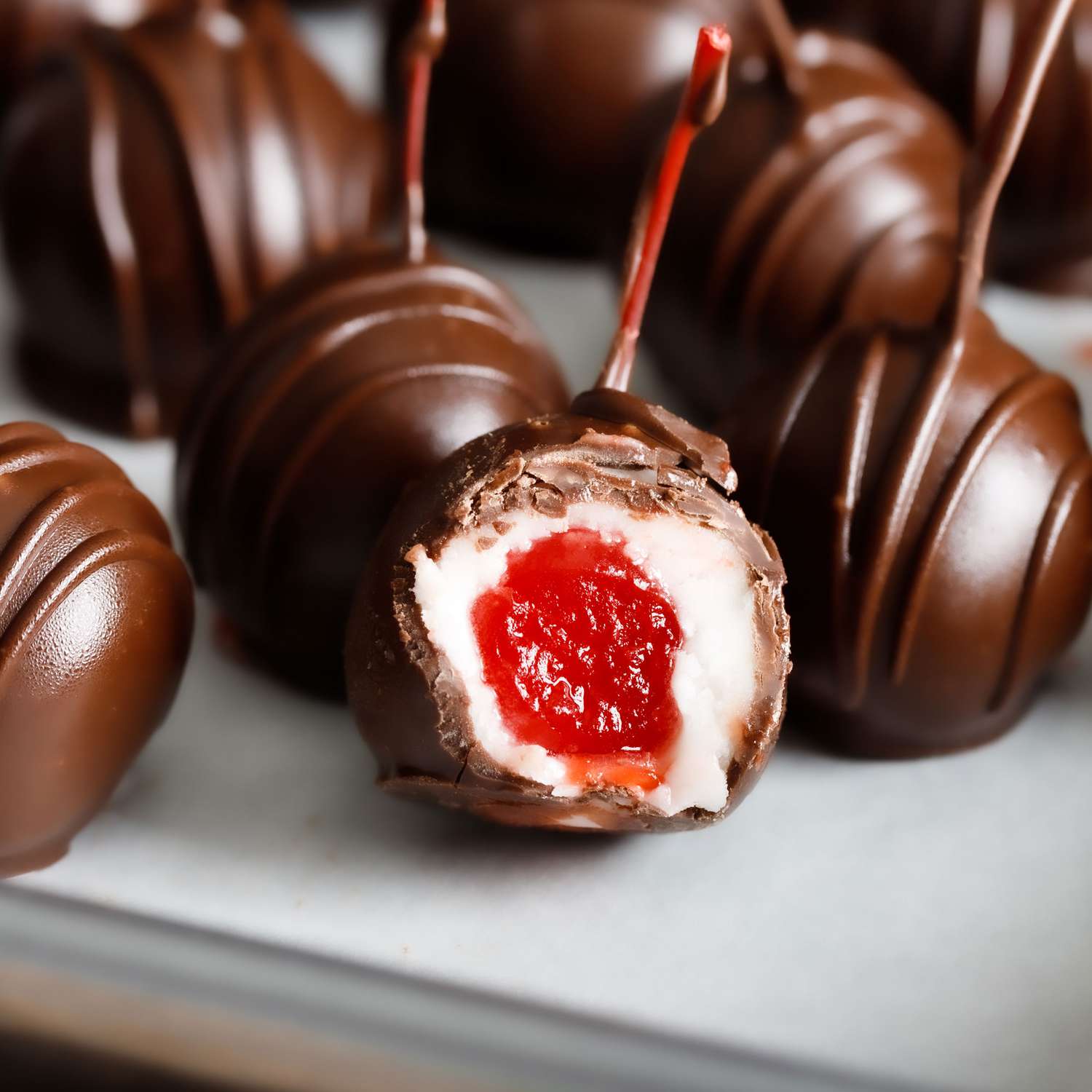 a close up view of several chocolate covered cherries chilling on a parchment lined baking sheet with one cut open revealing the creamy center wrapped around a bright red cherry.