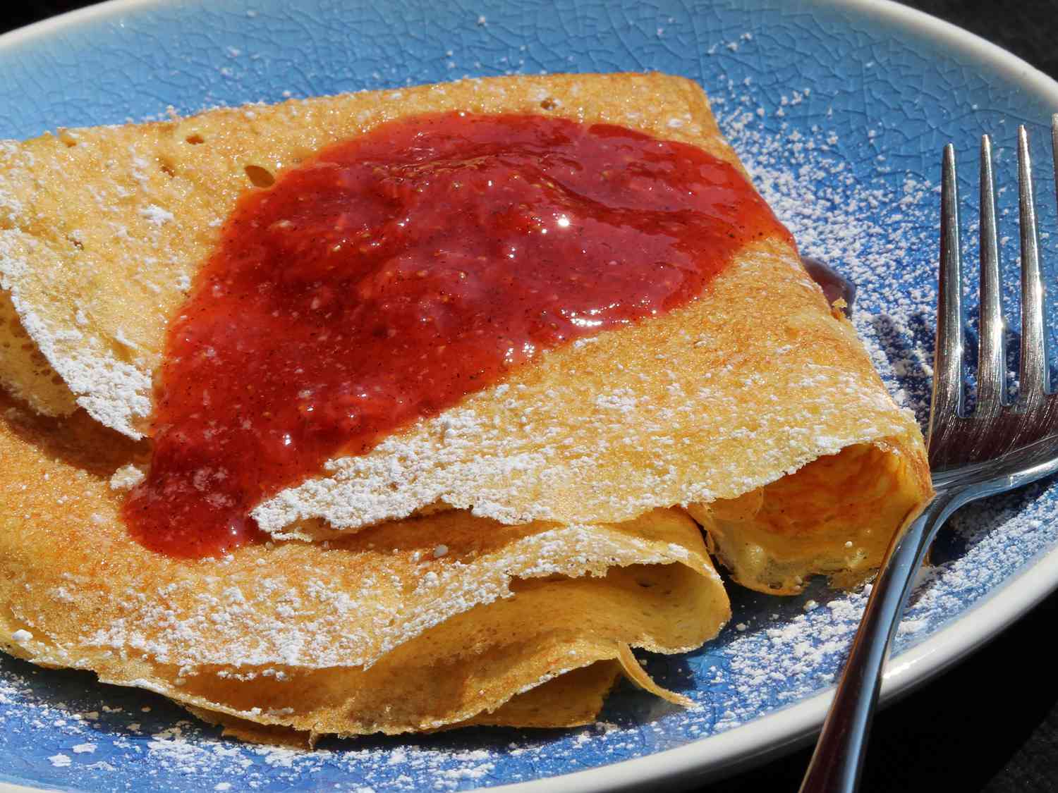 close up view of Finnish Pancakes garnished with jam and powdered sugar on a blue plate with a fork