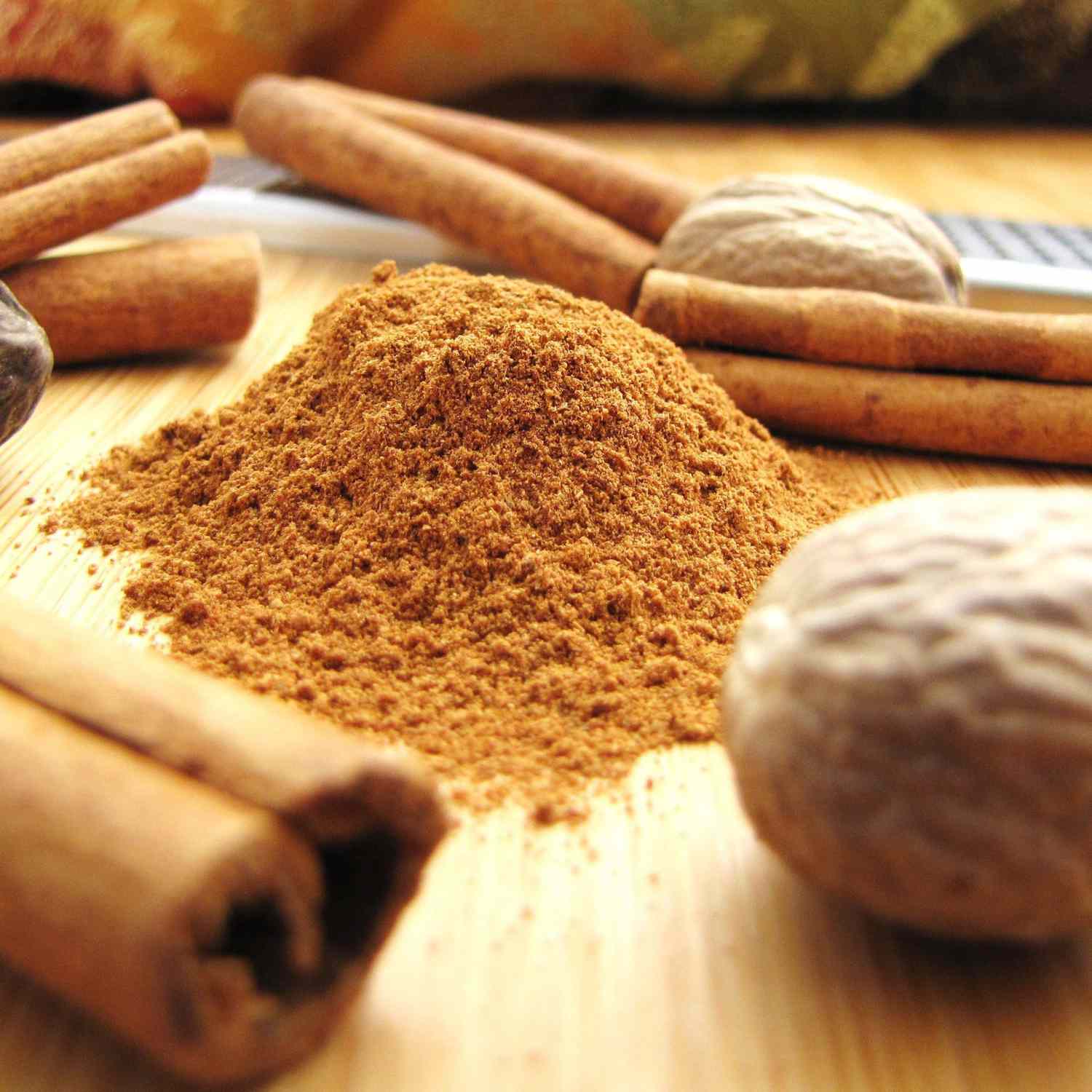 side view closeup of a mound of ground spices on a cutting board surrounded by whole nutmegs and cinnamon sticks, with a microplane in the background