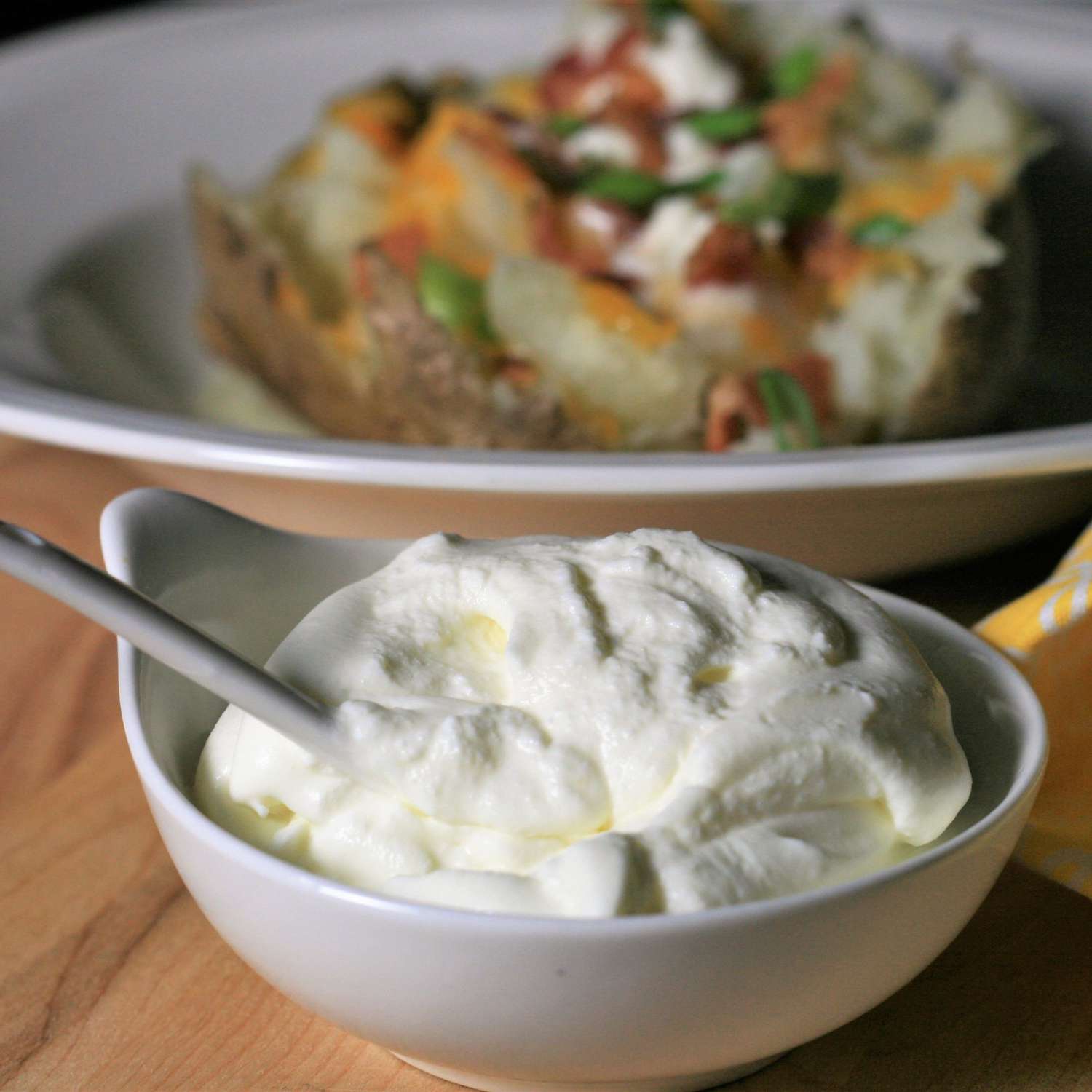 a spoon and serving bowl of thick-looking sour cream in the foreground with a loaded baked potato in the background