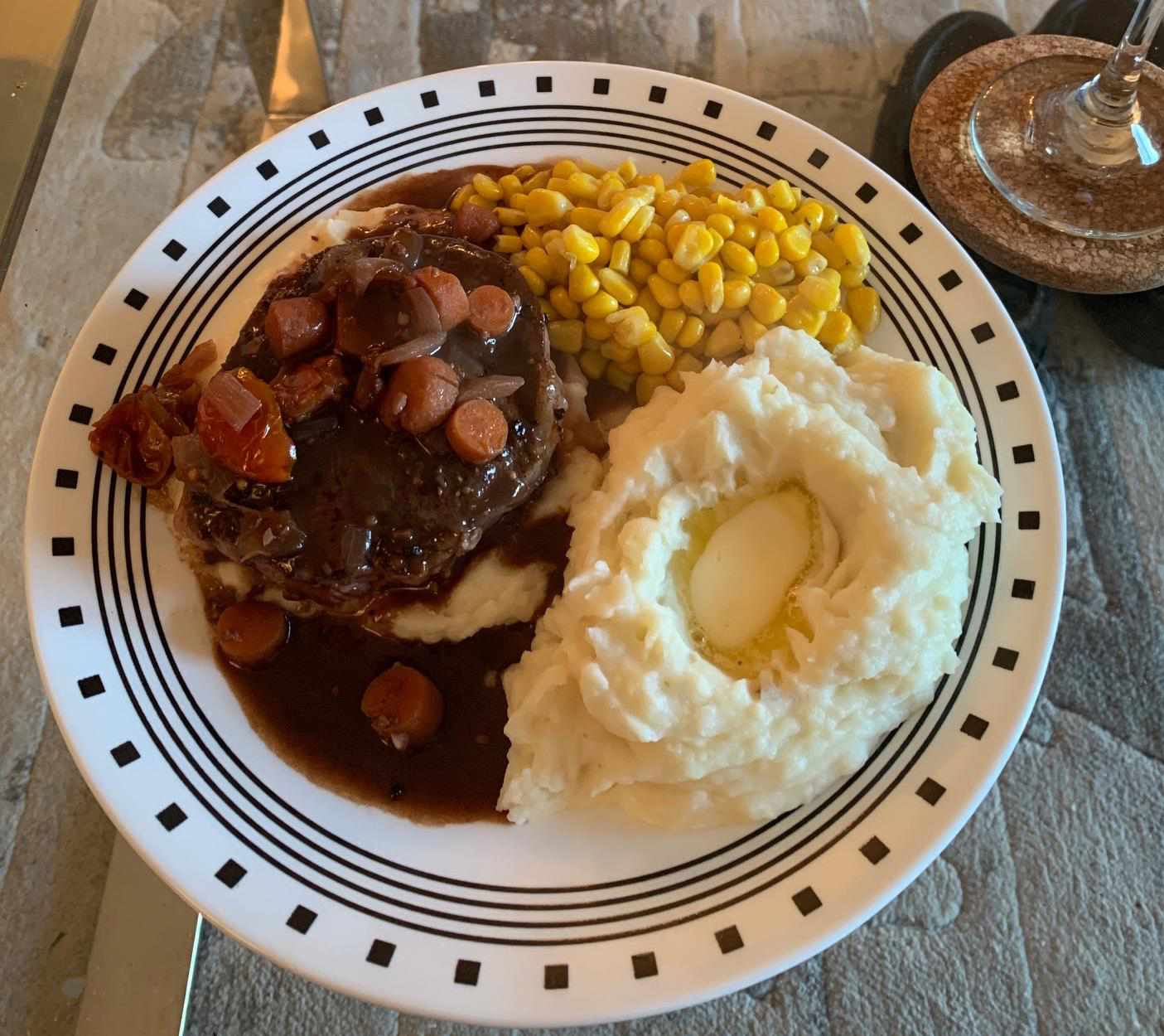 top-down view of a dinner plate with steak topped with sauce and vegetables with buttery mashed potatoes and corn on the side