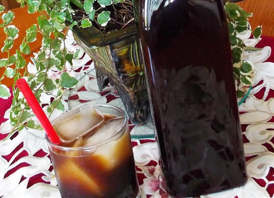 a glass of a dark brown drink with ice and a red straw, next to a tall bottle on a white cutwork placemat with a houseplant in the background