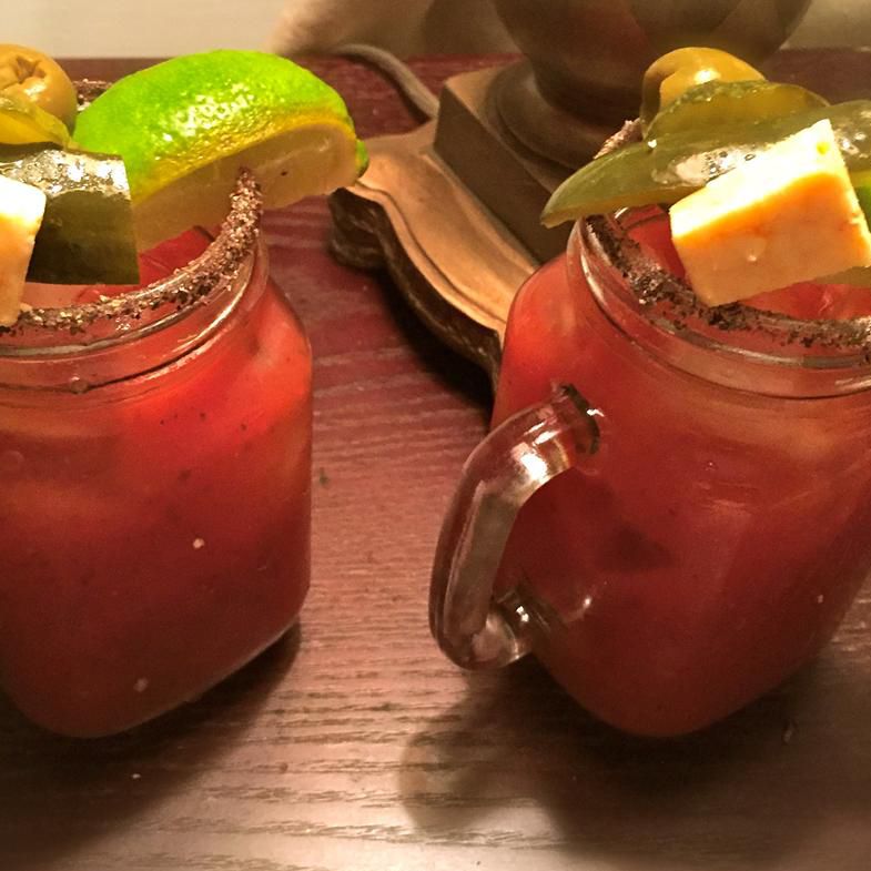 two mason jar mugs with seasoned rims holding bloody maryes garnished with lime wedges, pickle spears, olives, and a square of cheese