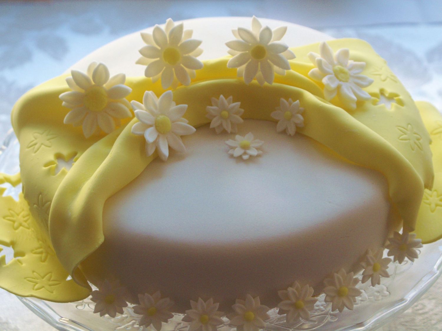 a cake covered with a smooth layer of white fondant with a yellow fondant ribbon and daisies