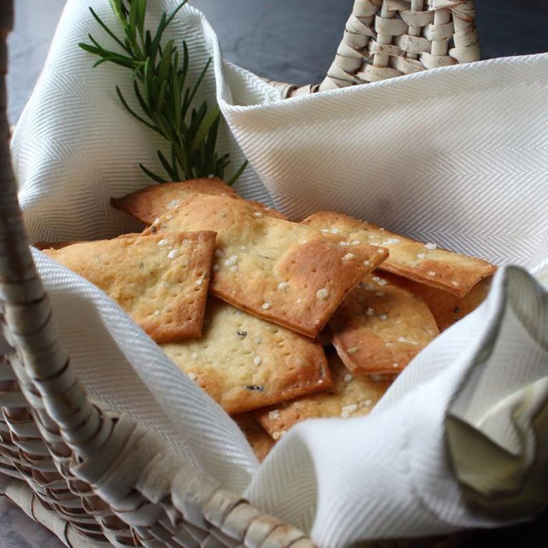 close up view of Crispy Rosemary Sea Salt Flatbread Crackers in a fabric lined basked to fresh herbs