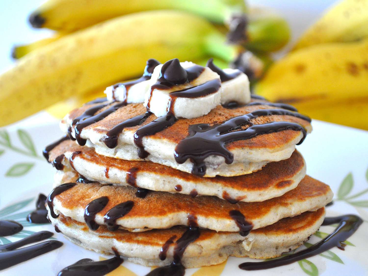close up view of Chunky Monkey Pancakes garnished with chocolate and bananas on a white plate, with bananas in the background