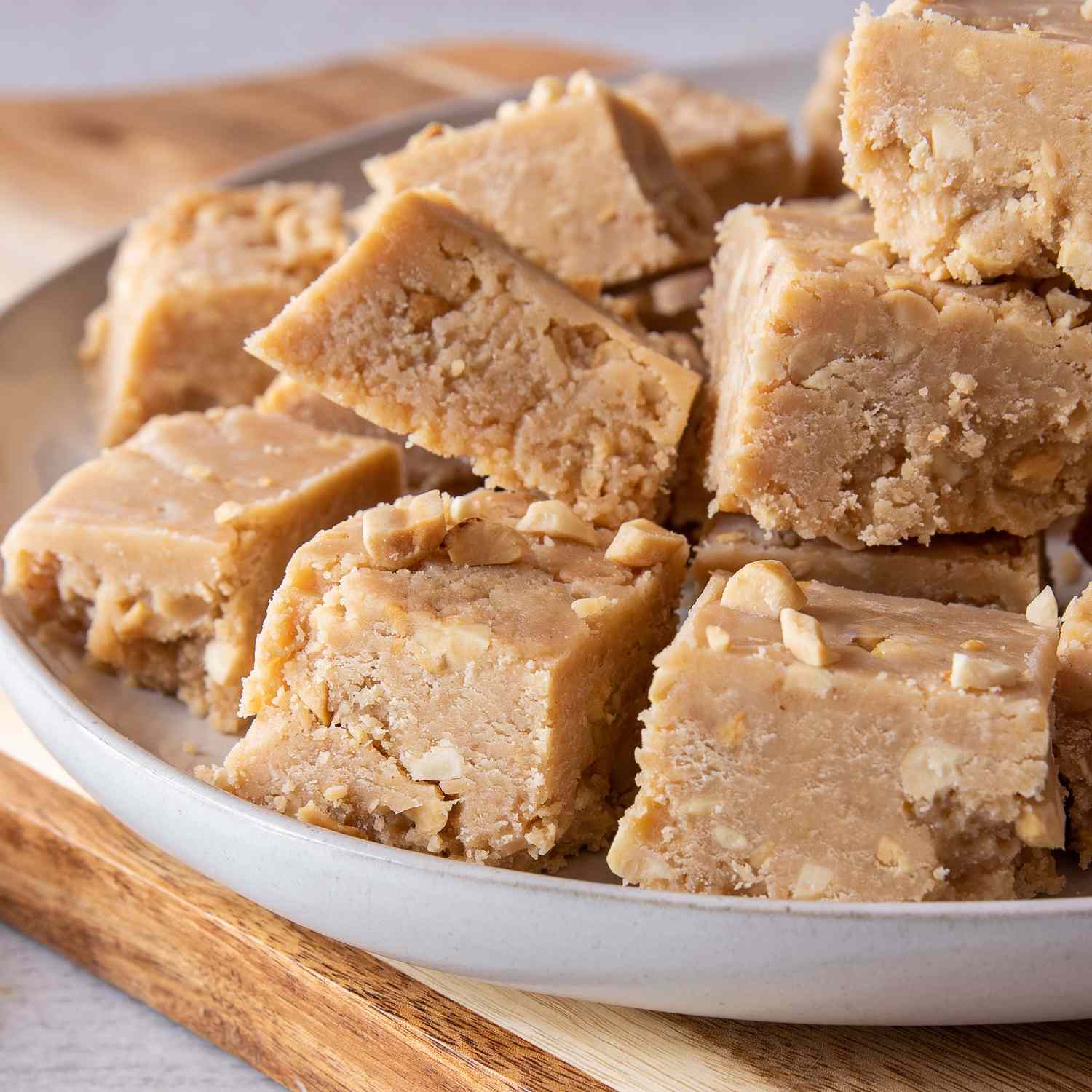 a low angle, close up view of a pile of peanut butter fudge squares stacked on a plate with a bowl of crushed peanuts sitting nearby.