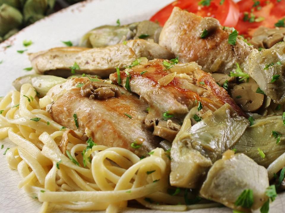 close up view of Chicken with Artichokes and Mushrooms over pasta, served with fresh herbs and tomatoes on a white plate
