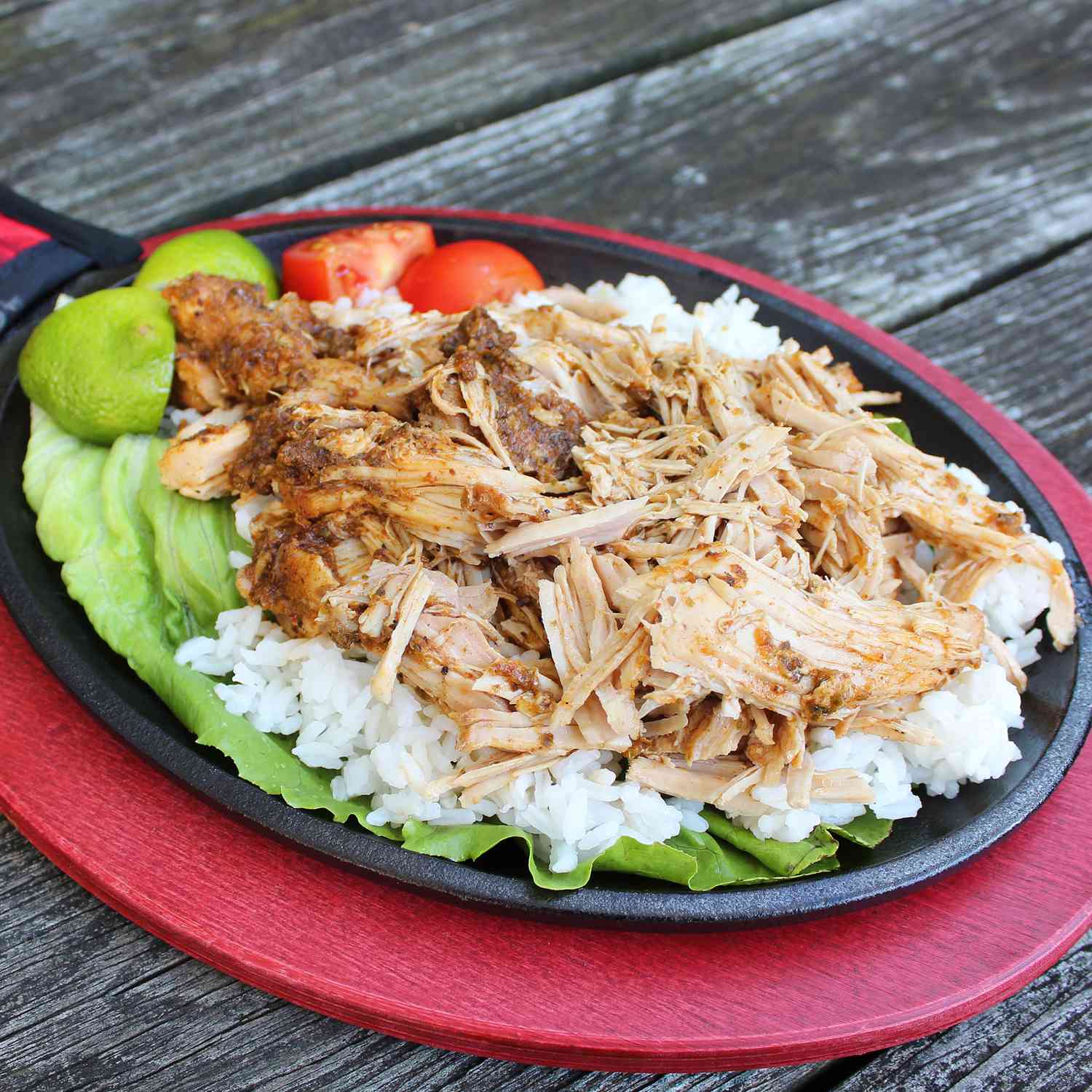 close up view of Slow Cooker Pernil Pork over white rice and lettuce, with lime wedges and tomatoes on a cast iron skillet