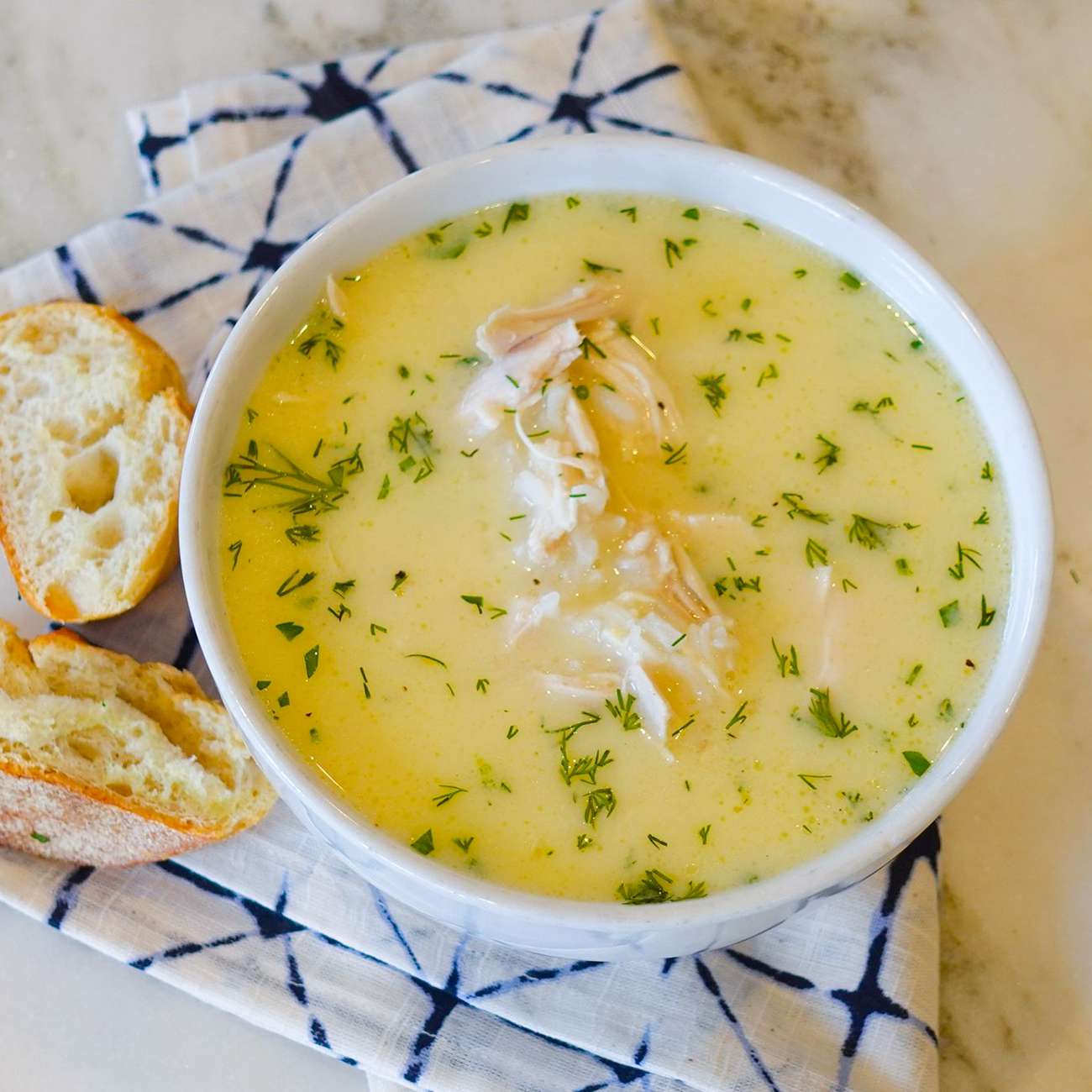 close up view of Greek Avgolemono Chicken Soup garnished with fresh herbs in a white bowl, served with bread