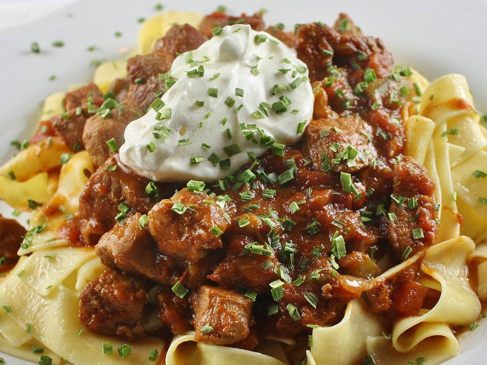close up view of Porkolt (Hungarian Stew) Made With Pork garnished with sour cream and herbs over pasta on a plate