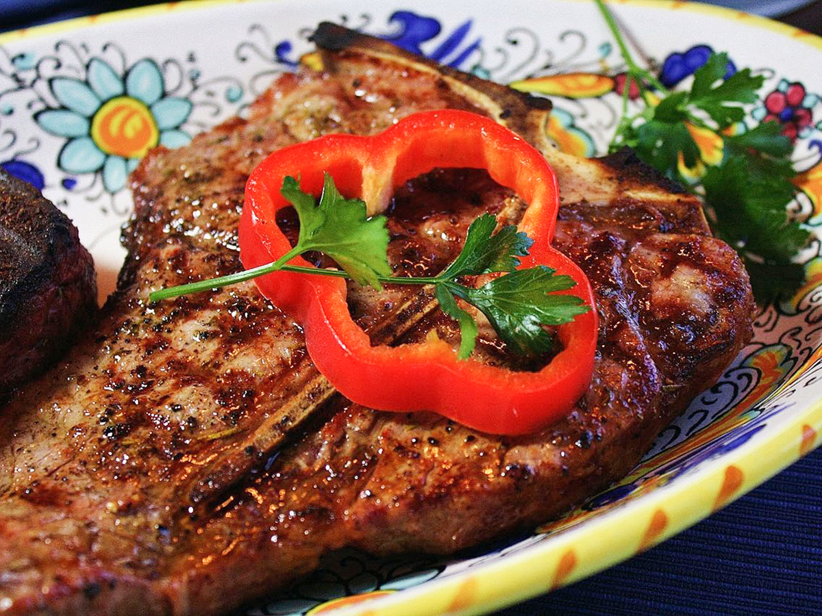 close up view of Bistecca alla Fiorentina with a red pepper sliced and fresh herbs on a colorful plate
