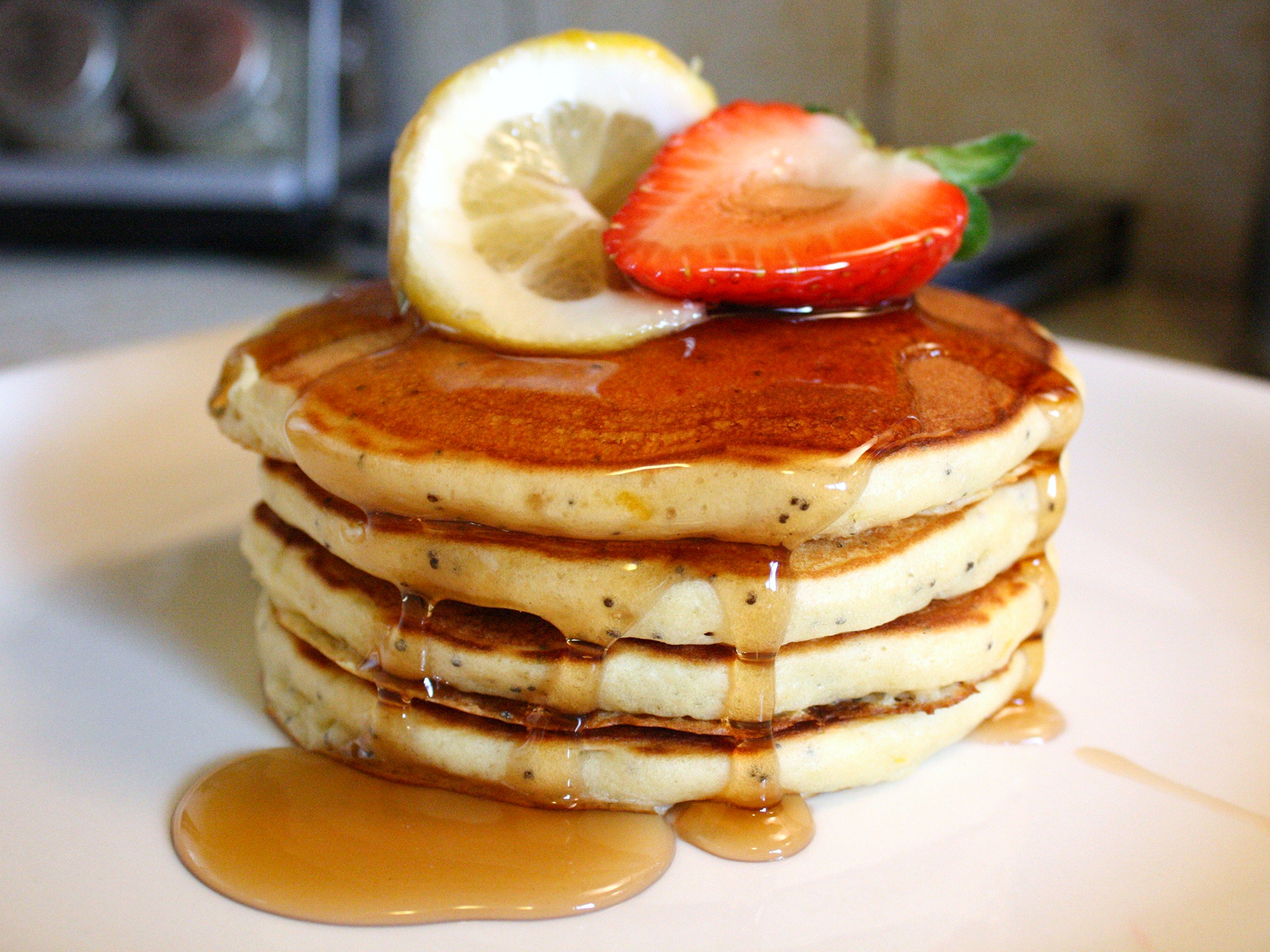 close up view of Sunday Morning Lemon Poppy Seed Pancakes garnished with syrup, strawberry and lemon on a plate