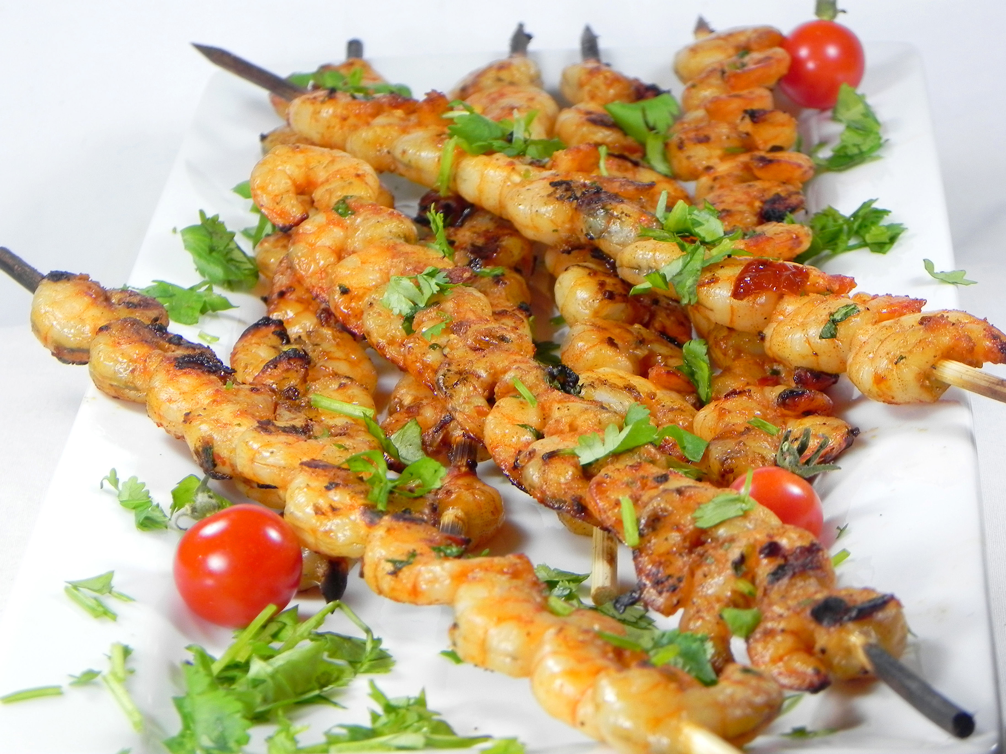 close up view of Spicy Chipotle Grilled Shrimp on skewers garnished with fresh herbs and cherry tomatoes on a white plate