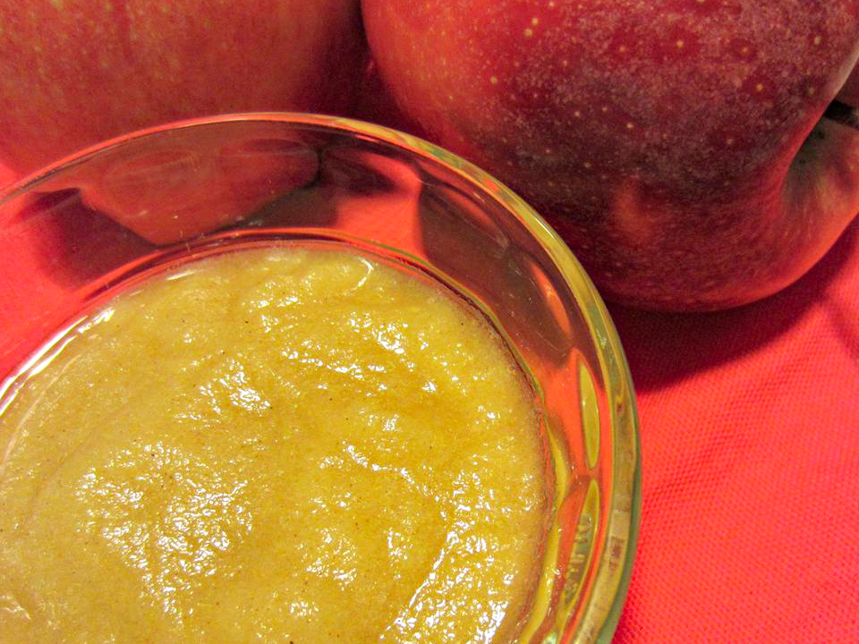 close up view of Applesauce in a glass bowl, with apples in the background, on a red surface
