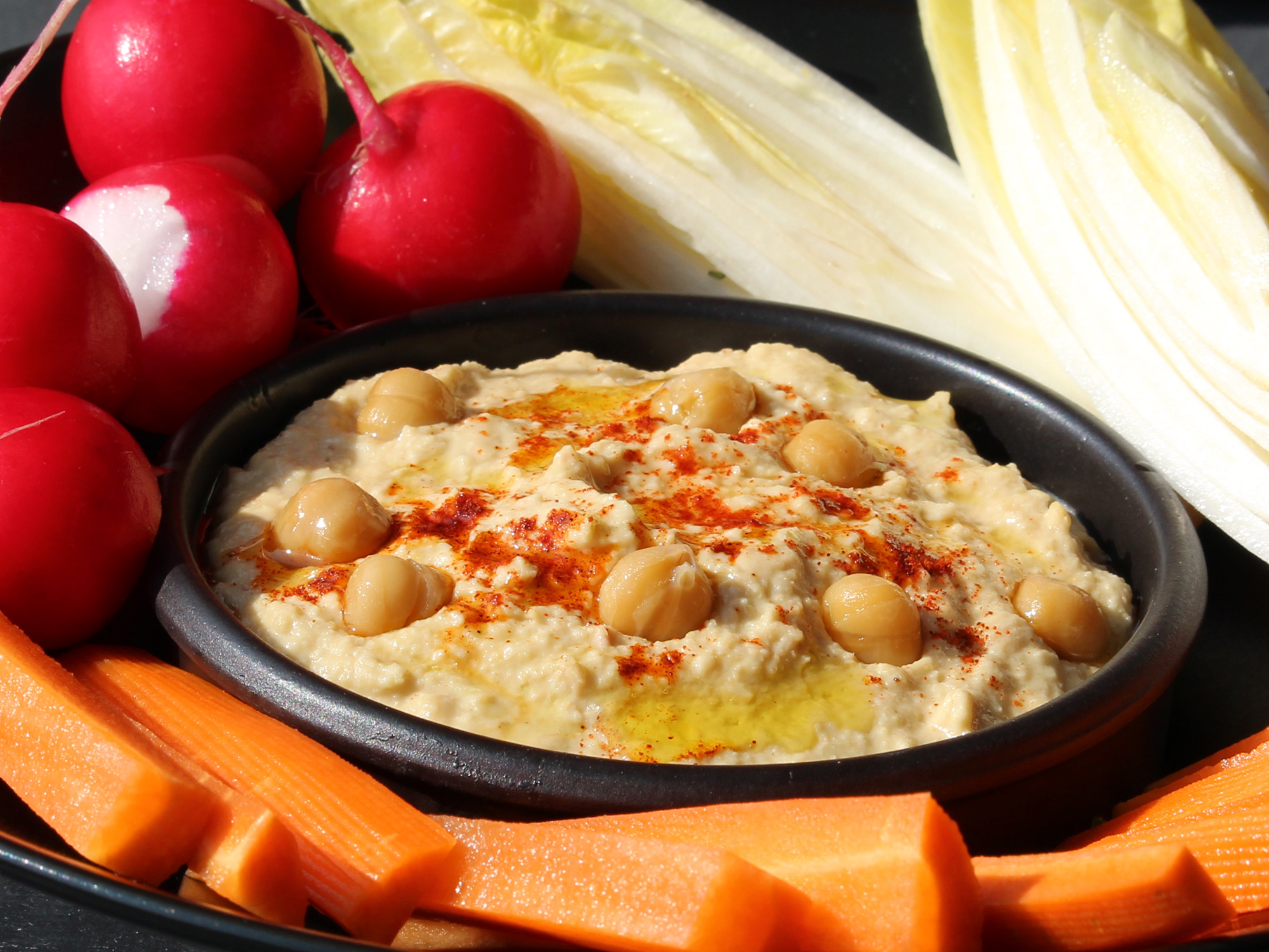 close up view of Middle Eastern Hummus (Chummus) in a black bowl, served with radishes, endives and carrots on a platter