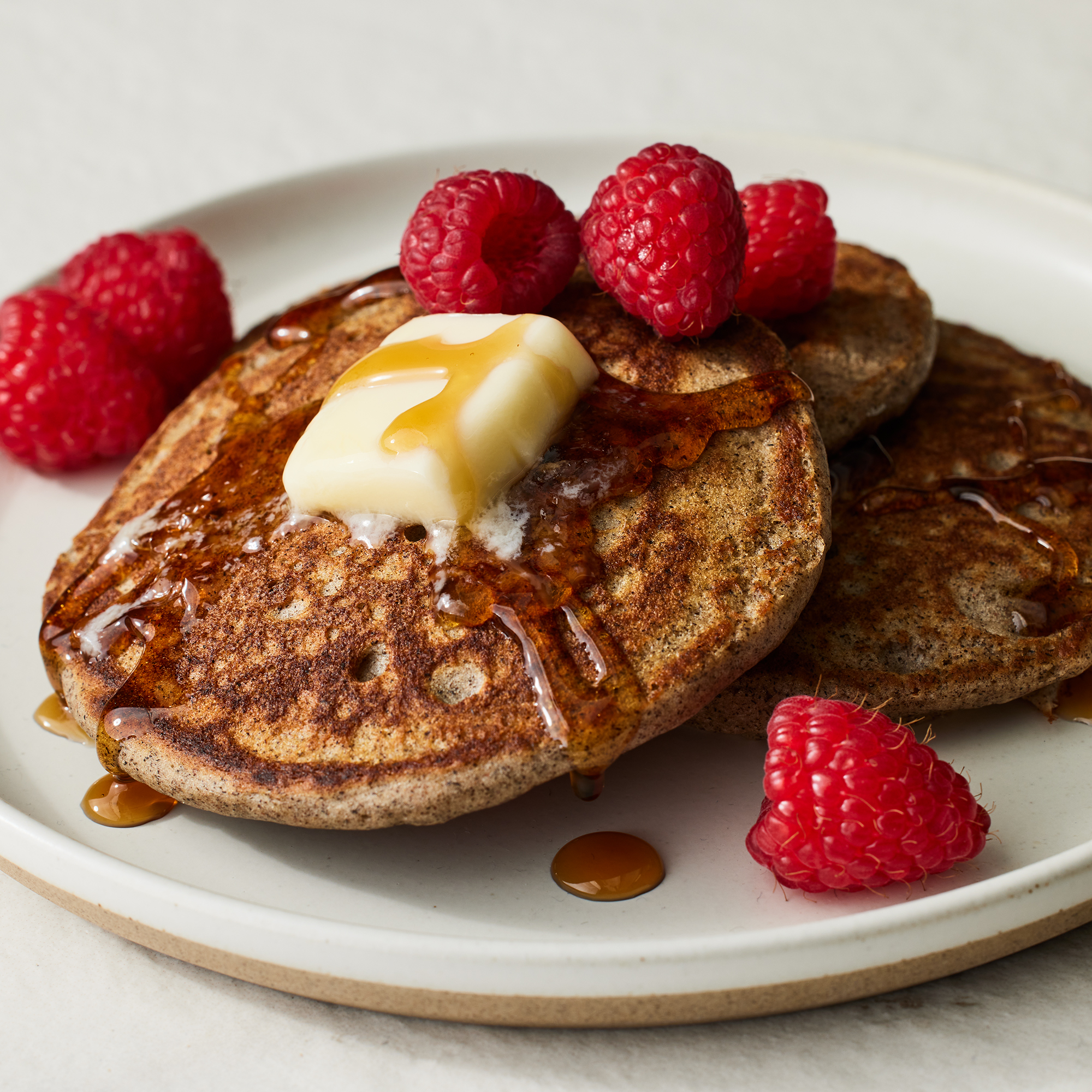 close up on a stack of buckwheat pancakes topped with butter, syrup and fresh raspberries