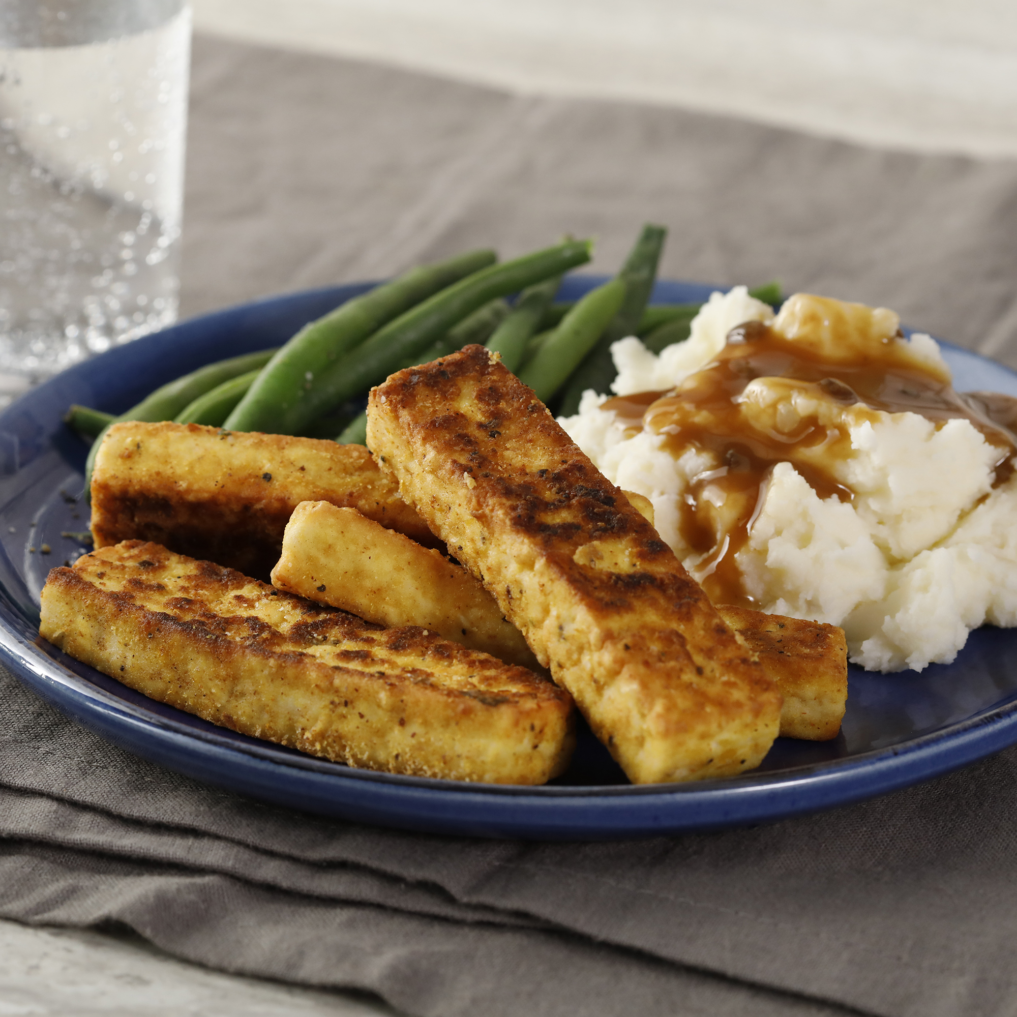 close up view of Breaded, Fried, Softly Spiced Tofu served with green beans and mashed potatoes and gravy on a blue plate