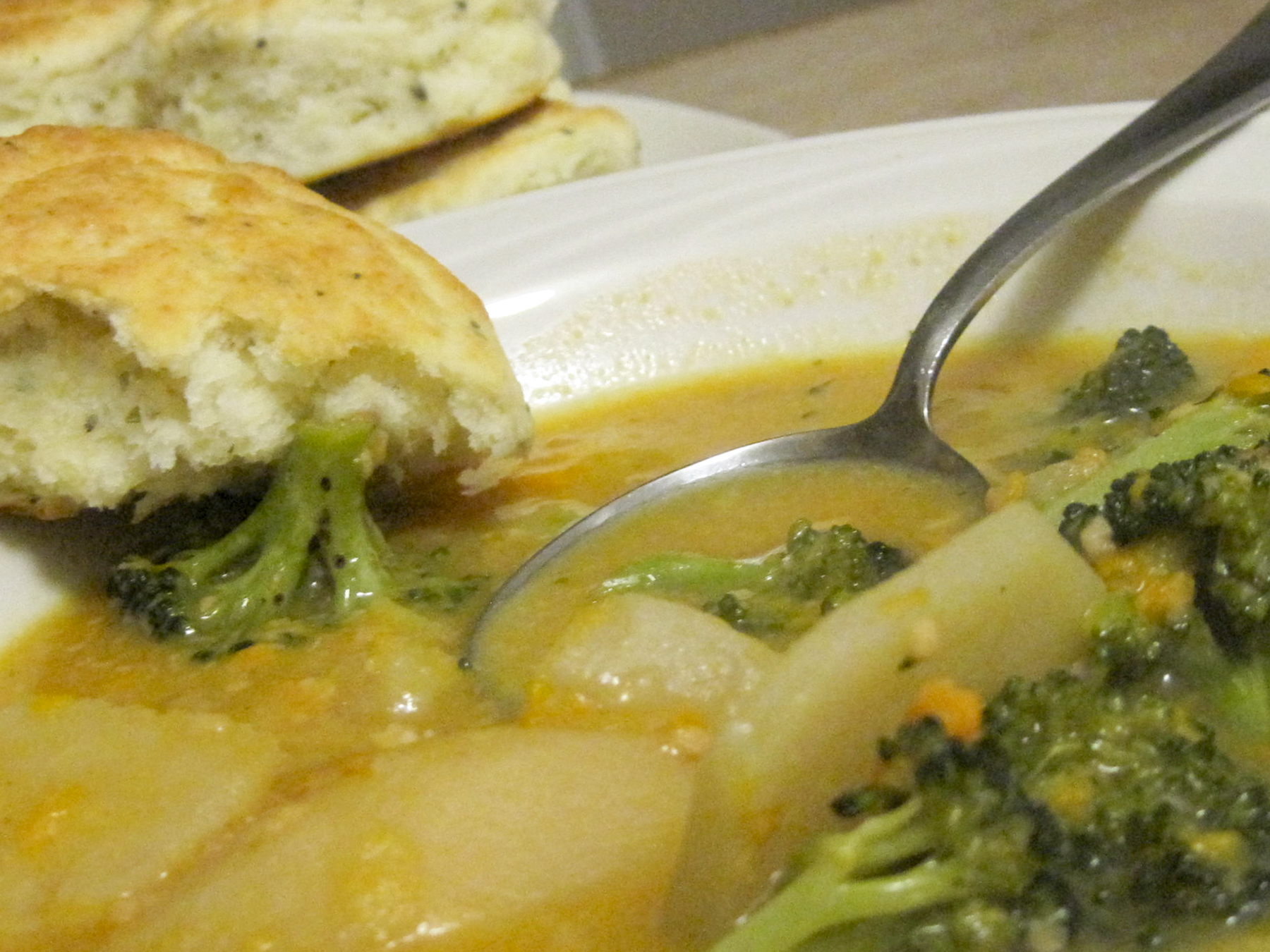 close up view of Scalloped Potato Soup with broccoli in a white bowl with a spoon, with biscuits