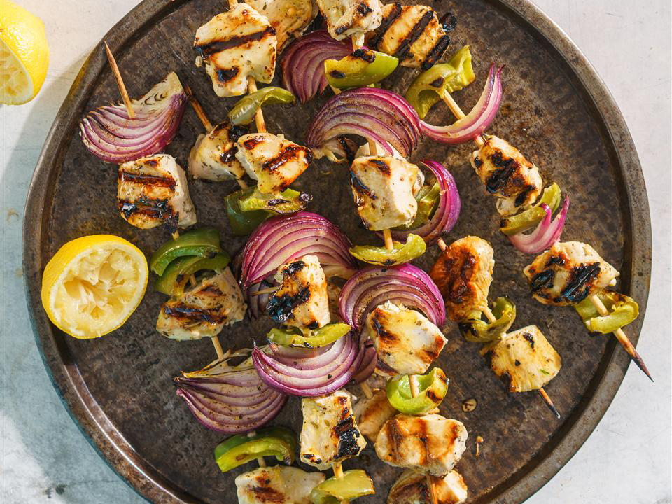 close up view of Marinated Greek Chicken Kabobs with red onions and green onions on wooden skewers, on wooden skewers on a platter with a lemon